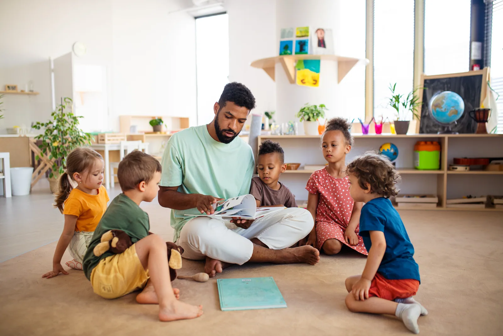 A teacher sitting cross-legged on the floor reading a book to a circle of children