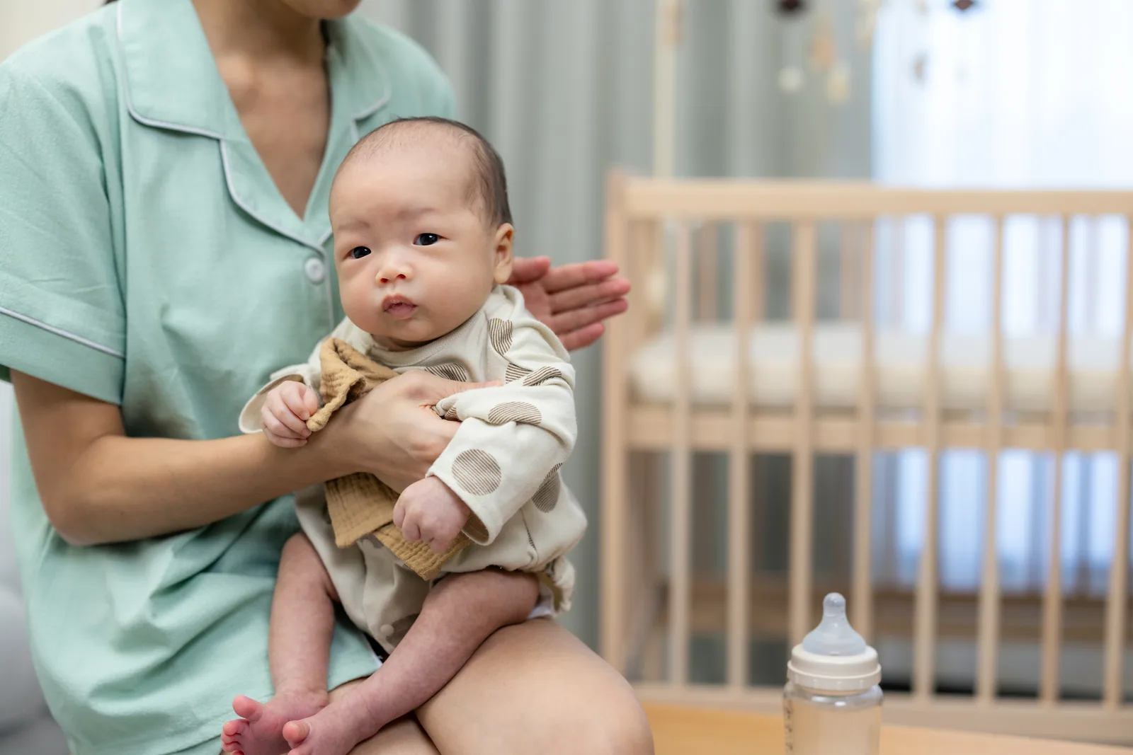 A caregiver sits beside a crib holding an infant in a quiet, softly lit room