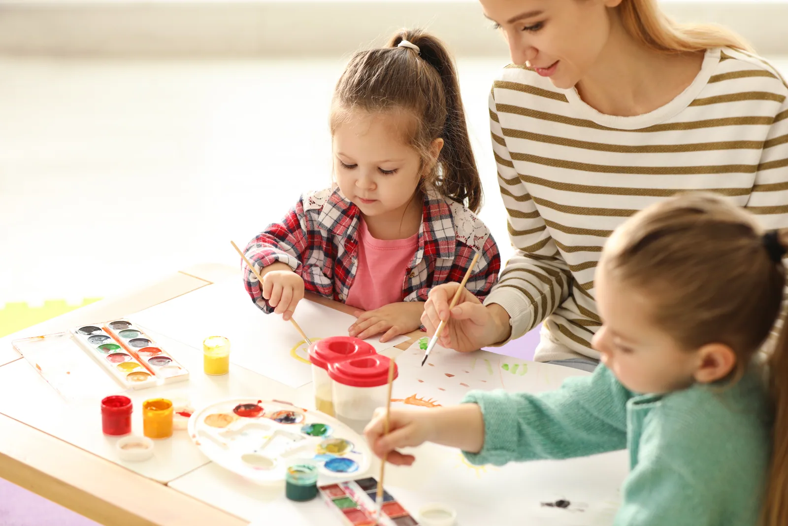 A teacher and two young children painting at a wooden table in warm daylight