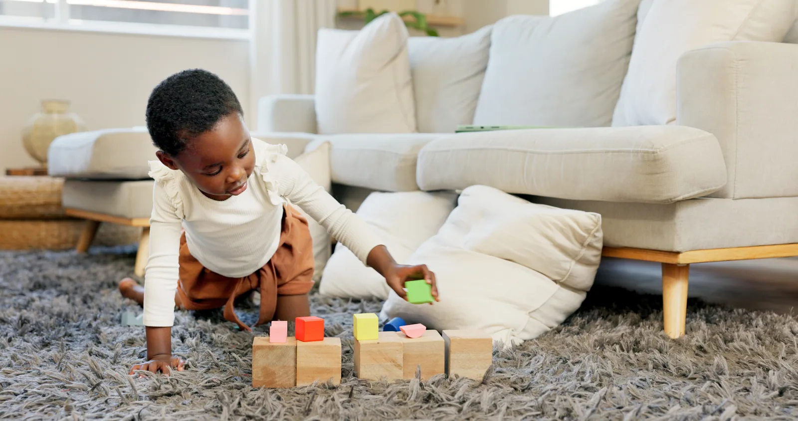 A toddler kneels on a rug, reaching for a stack of wooden and colored blocks